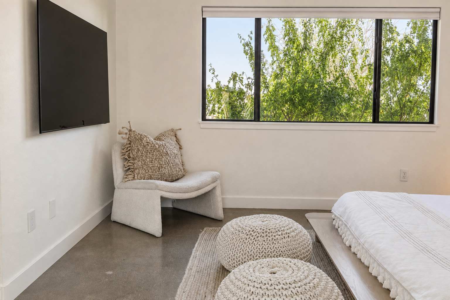 A bedroom with a bed, a seating area with cushions, a large window with a view of trees, and a flat-screen TV mounted on the wall.