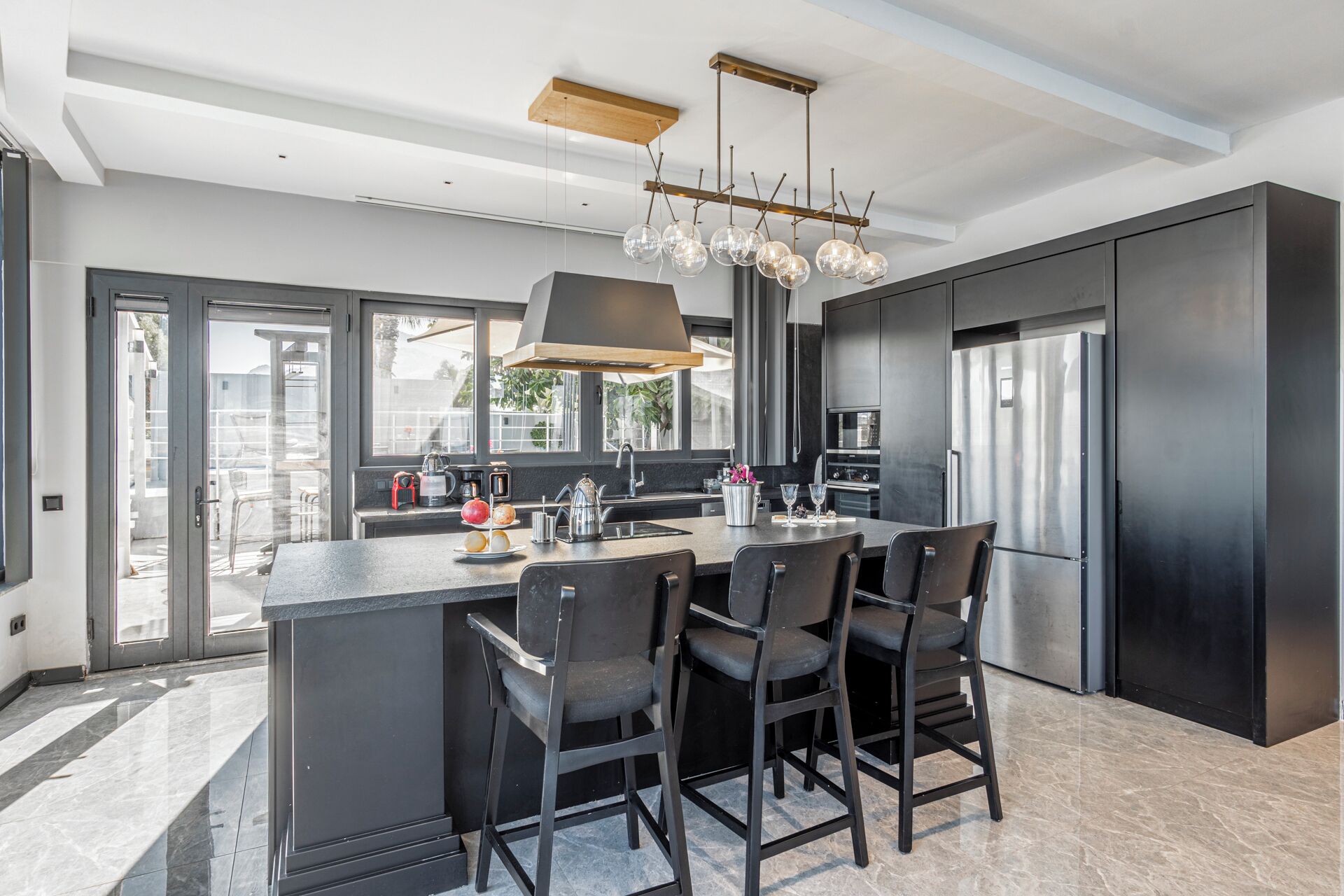 A modern kitchen with a central island, black chairs, a stainless steel refrigerator, and a large window providing natural light.