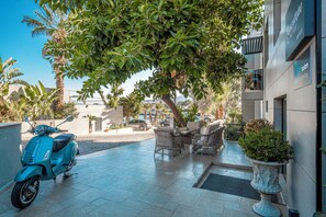 A modern outdoor seating area with a blue scooter, wicker furniture, and a large tree providing shade. - Çimentepe Residence Deluxe. (Bodrum)