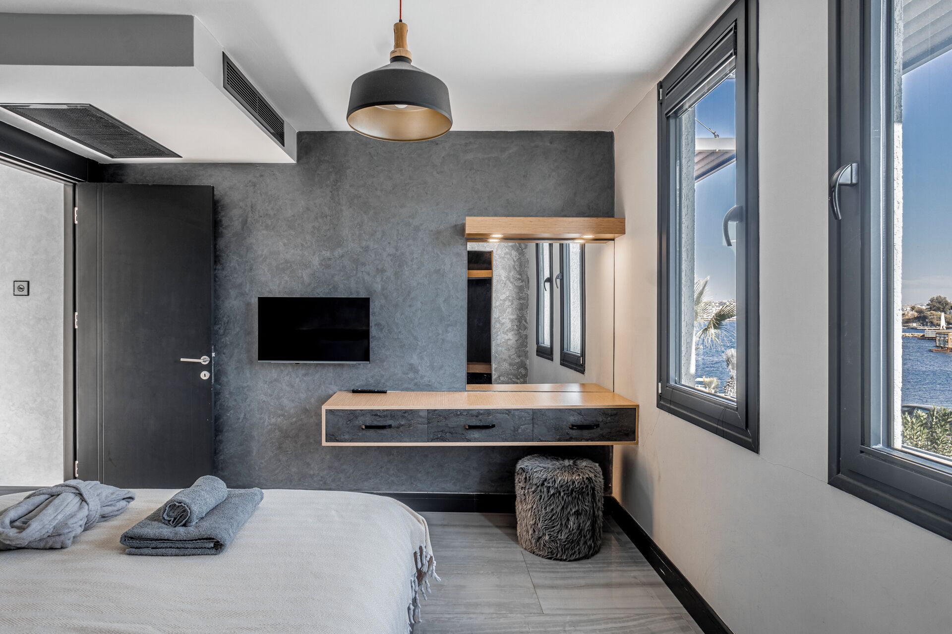 A modern bedroom with a large window, a flat-screen TV, a wooden bedside table, and a built-in bench.