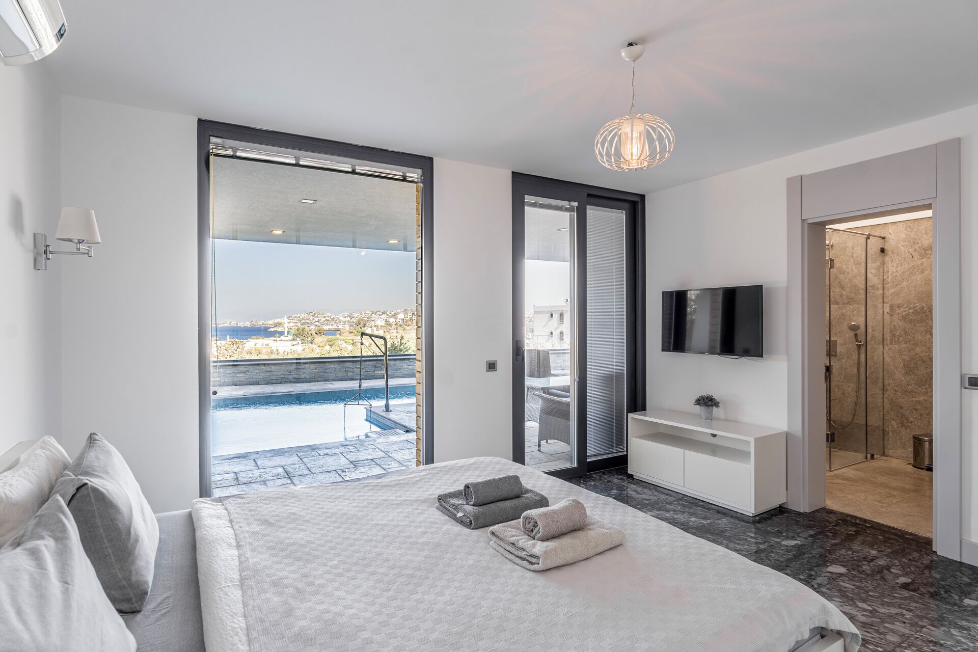 A modern bedroom with a large bed, a TV, a bathroom with a shower, and a balcony with a city view.