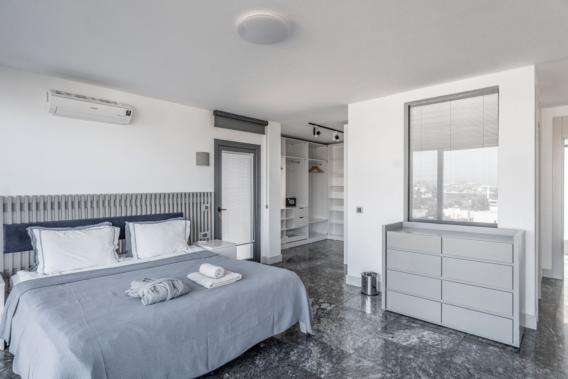 Ein modernes Schlafzimmer mit Bett, Nachttischen, begehbarem Kleiderschrank und einem Fenster mit Jalousien.