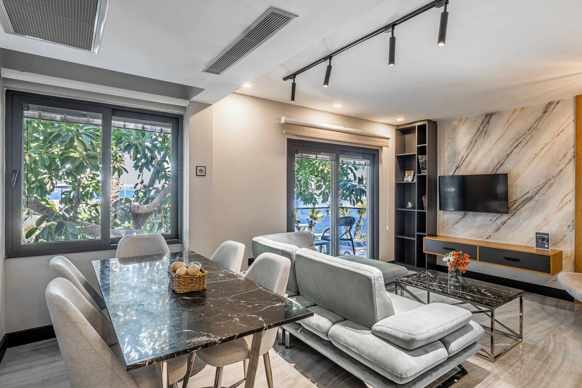 Un salon moderne comprenant un coin repas, de grandes fenêtres, un téléviseur à écran plat et un mur décoré d’un motif imitant le marbre.