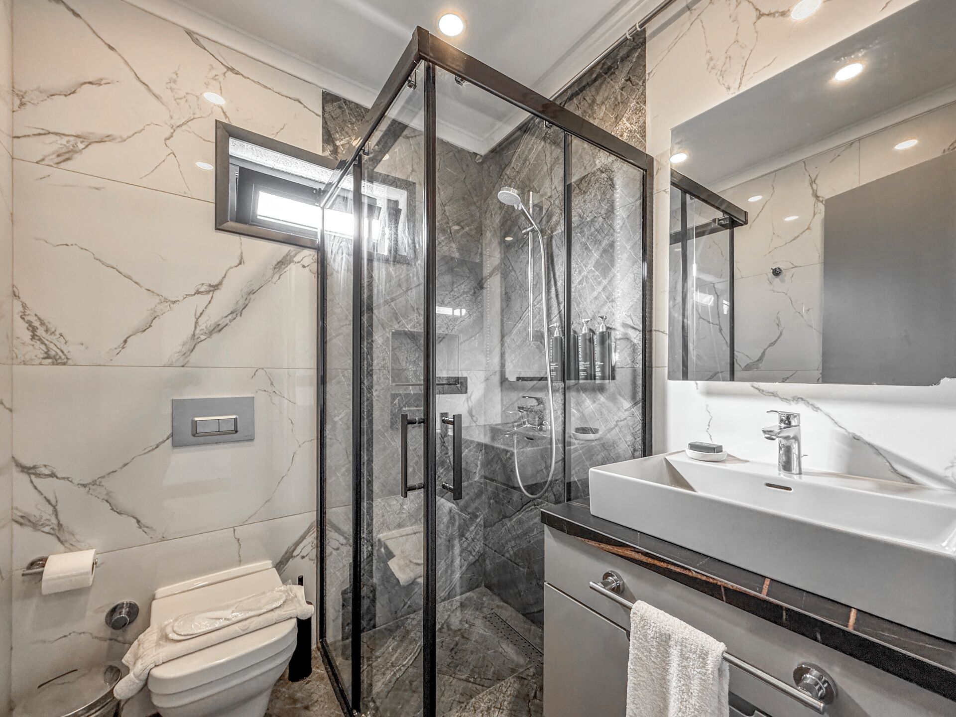 A modern bathroom with a glass-enclosed shower, a marble-tiled wall, a white sink, and a large mirror.