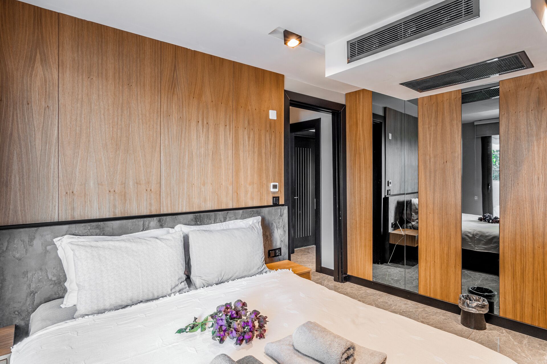 A modern bedroom with a large bed, wooden panel walls, a mirror, and a visible bathroom.