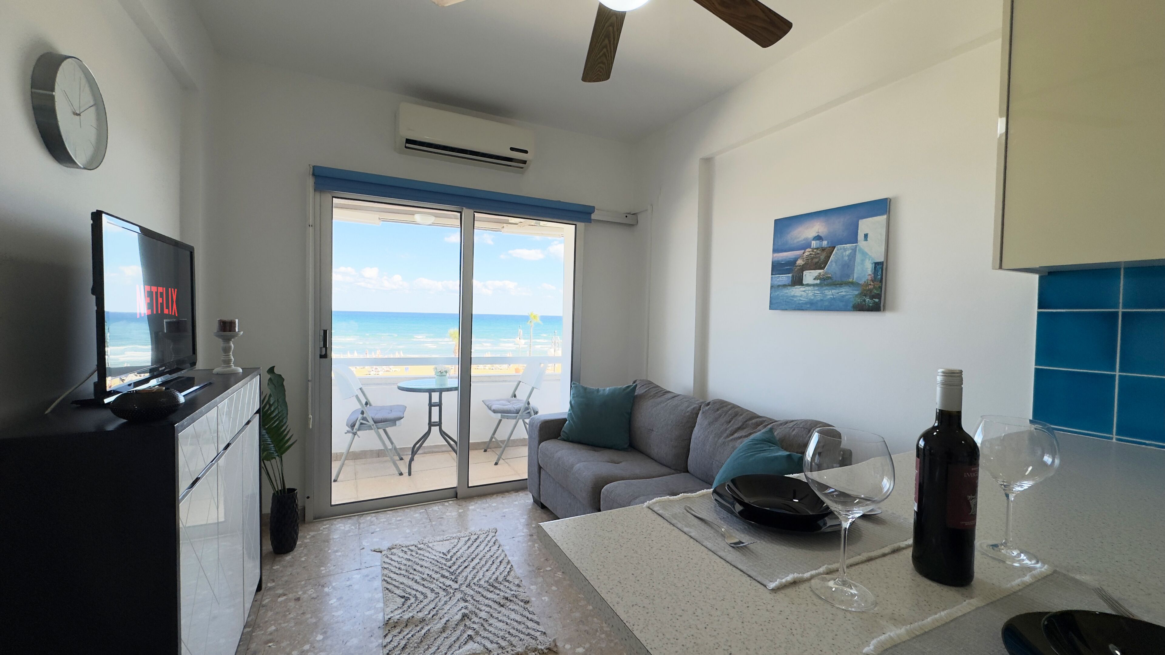 Uma sala de estar moderna com uma TV exibindo 'NETFLIX', uma varanda com vista para a praia e uma área de jantar com uma garrafa de vinho e um copo.