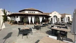 Un espace extérieur aménagé en terrasse avec des tables et des chaises, un coin salon couvert avec des rideaux, et un bâtiment doté d’un toit en tuiles.