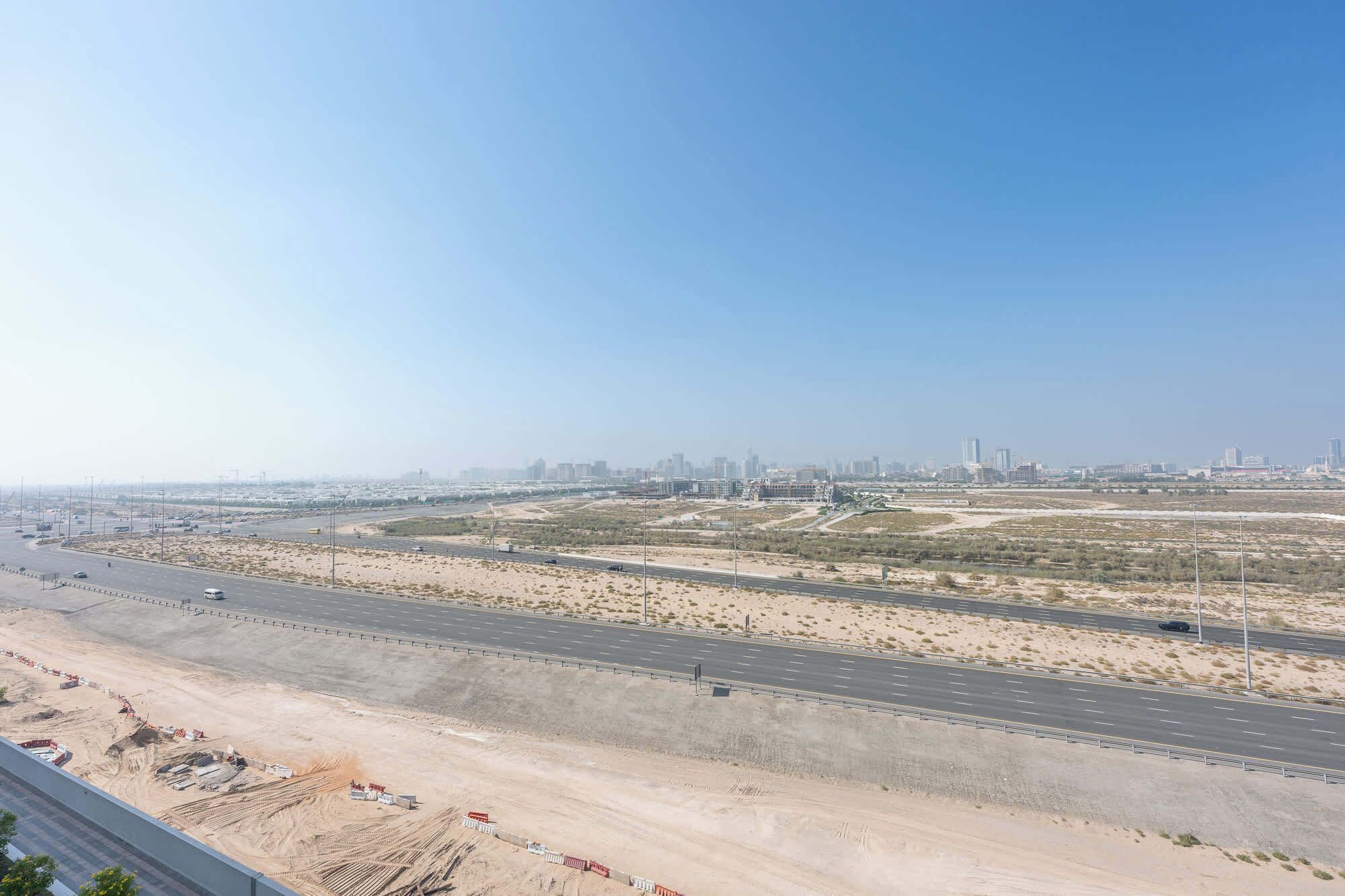 Autostrada in costruzione, paesaggio desertico e edifici cittadini in lontananza.