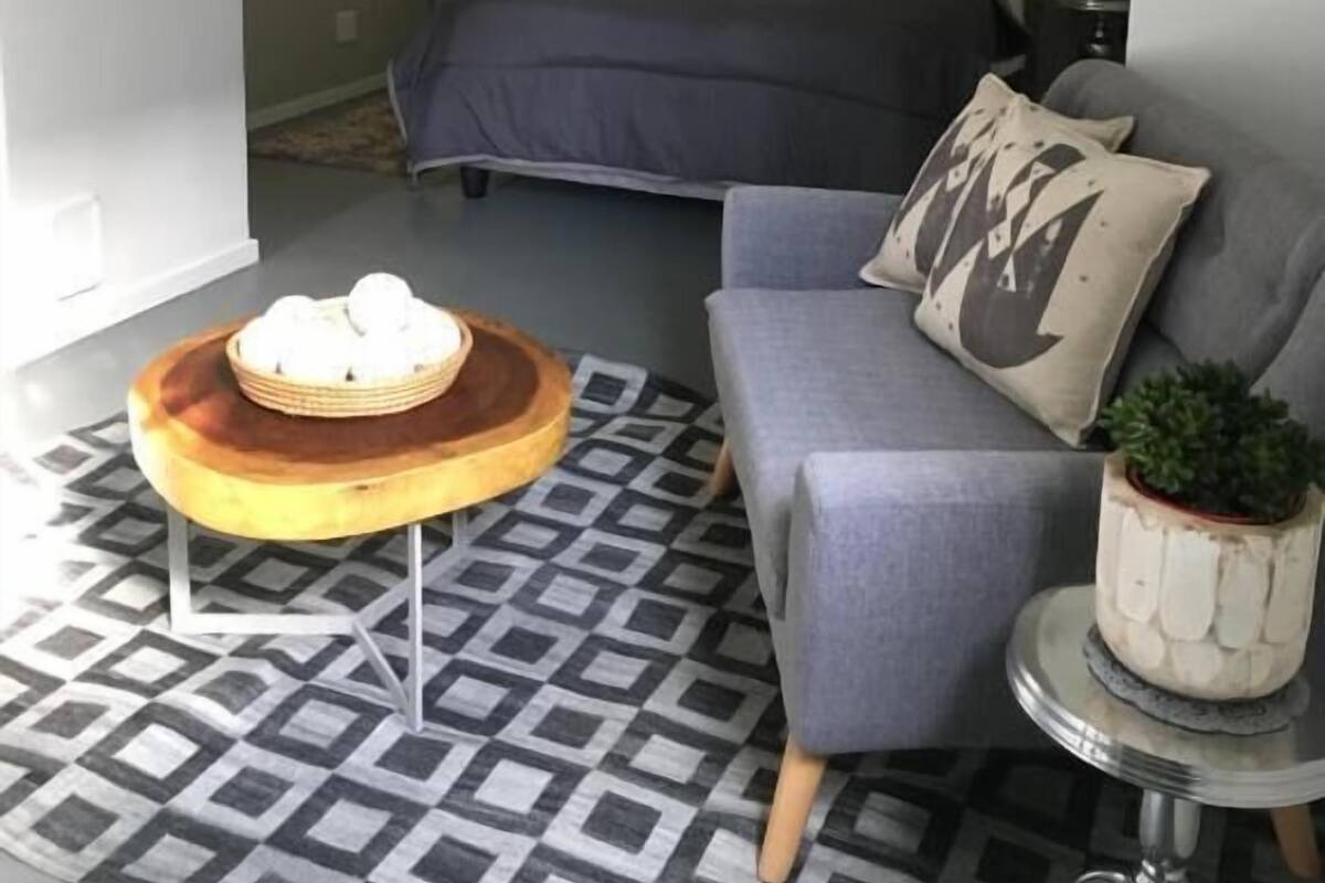 Um quarto moderno com uma cama, um sofá cinza, uma mesinha redonda com um cesto e um tapete estampado.