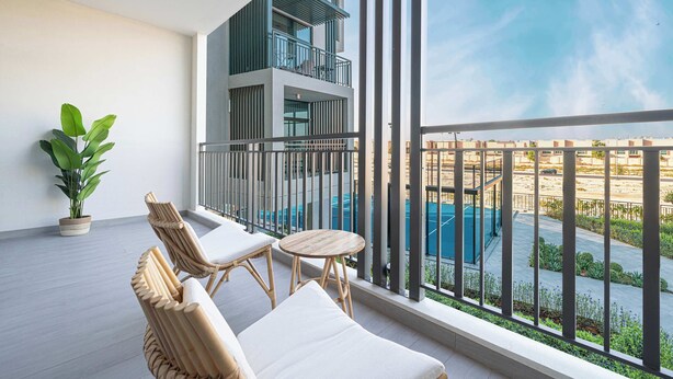 A balcony with wicker furniture, a potted plant, and a view of a swimming pool and buildings. - Modern 1-Bedroom Apartment With Padel Court Access (Arjan)
