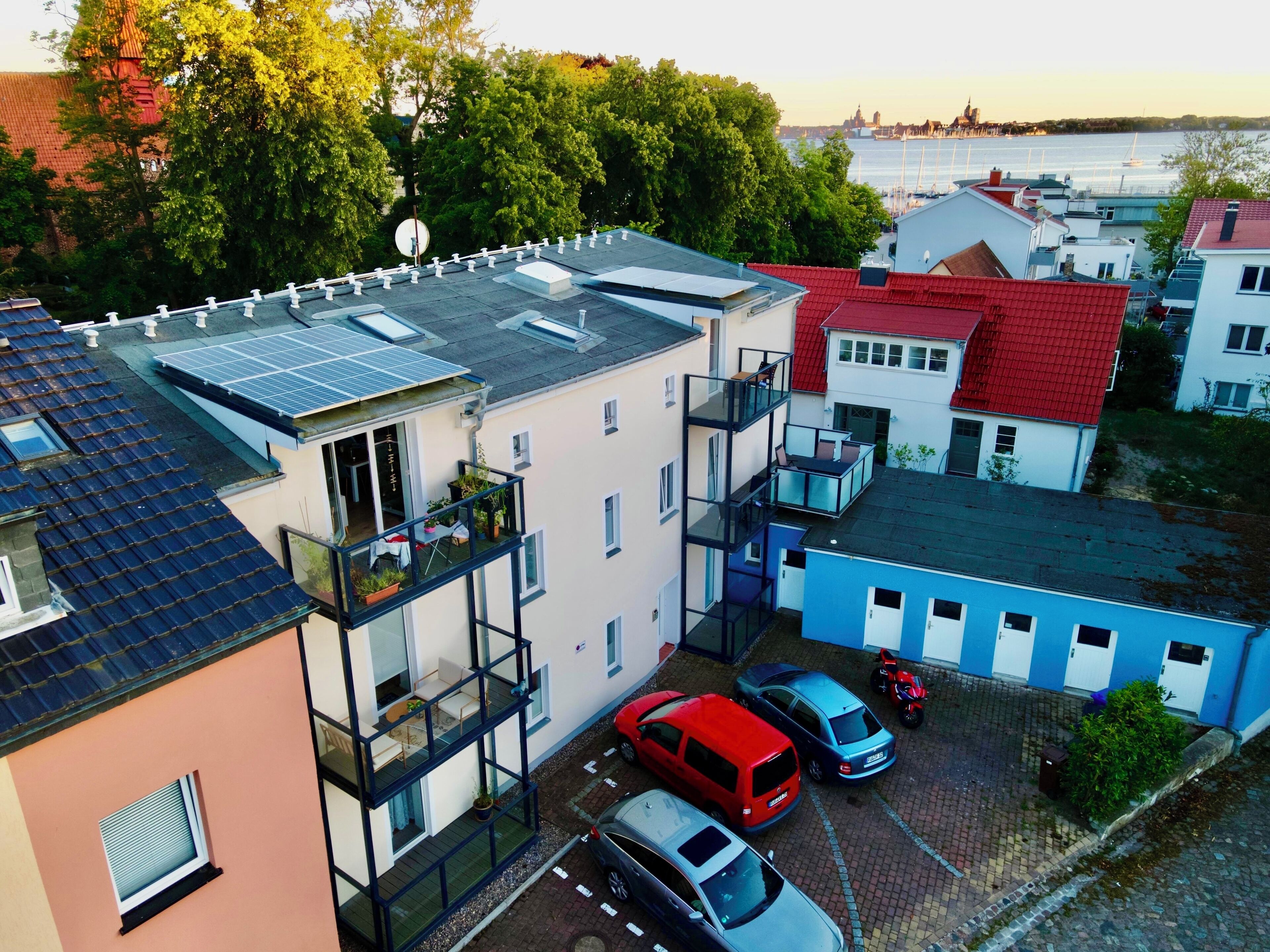 A multi-story residential building with balconies, solar panels, and a parking area with cars.
