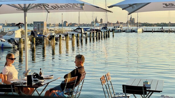 A waterfront restaurant with outdoor seating, a view of the marina, and a city skyline in the background. - Living with a harbor view—200 meters from the South Beach on the island of Rügen (Altefähr)