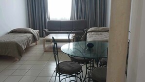 A room with two single beds, a sofa, a glass-top dining table with four chairs, and a window with curtains.