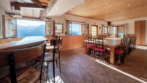 A spacious dining area with wooden tables and chairs, large windows with mountain views, and a high ceiling with wooden beams.