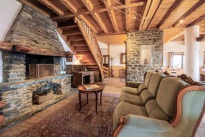 Ein gemütliches Wohnzimmer mit Kamin aus Stein, Holzbalken, einer Treppe und einer bequemen Couch.