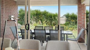 An outdoor patio with a glass door, metal chairs, a table, and a view of a garden with plants and a building in the background. - 15 Offshore Dr Middleton - Wi-Fi and Linen Included (Middleton)