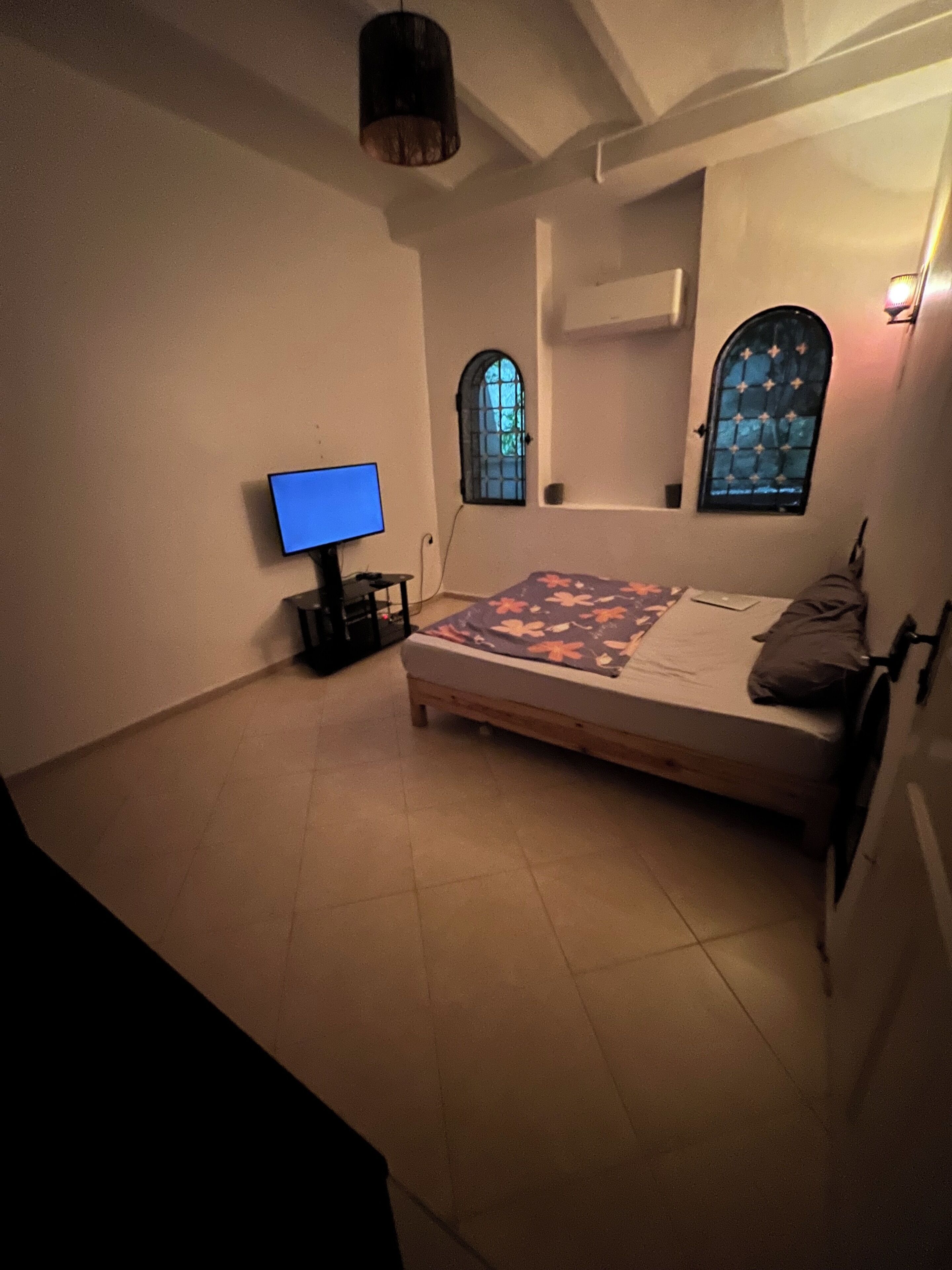 Une chambre à coucher comprenant un lit, un téléviseur posé sur un meuble, et deux fenêtres ornées de vitraux décoratifs.