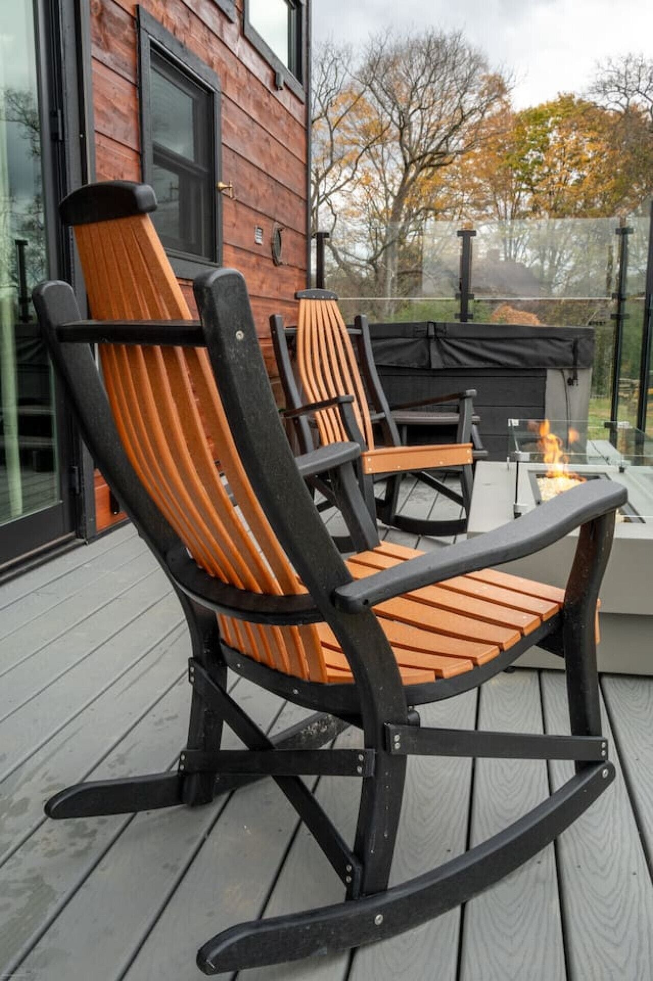 A wooden rocking chair and a table on a deck with a glass enclosure, overlooking a natural landscape.