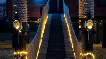 Un puente de madera con barandillas de cuerda y luces colgantes que lleva a un edificio con un letrero de neón.