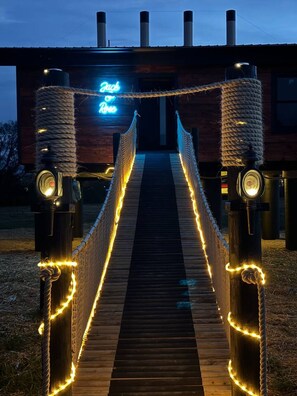 A wooden bridge with rope railings and string lights leading to a building with a neon sign. - Jack & Rose Treehouse Titanic Hot tub Riverfront. (Sevierville)