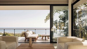 Een moderne buitenruimte met uitzicht op de oceaan, met een houten tafel, twee banken en een glazen deur naar een balkon.