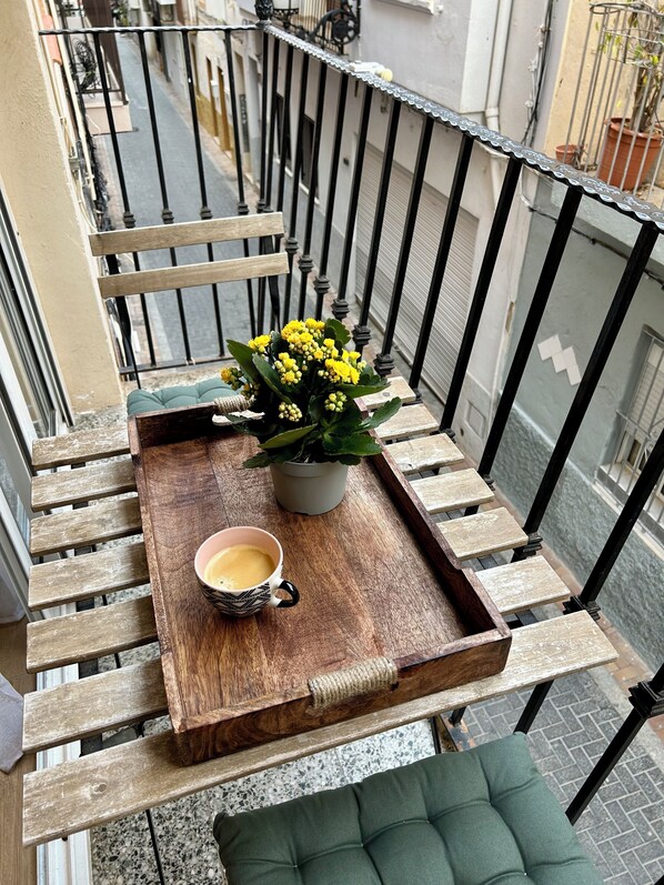 A balcony with a wooden bench, a potted plant with yellow flowers, and a tray with a cup of coffee.