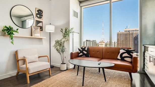 Ein modernes Wohnzimmer mit Sofa, Sessel und Couchtisch, großen Fenstern mit Stadtblick und minimalistischer Einrichtung.