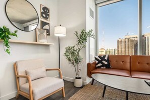 Un salon moderne comprenant un canapé, un fauteuil, une table ronde et une grande fenêtre donnant sur la ville.