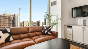 A modern living room with a leather sofa, a flat-screen TV, and a view of a cityscape through large windows. - Boulevard Stays Downtown Highrise STU Sky Pool, Parking, Gym 06 (Phoenix)