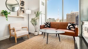 A modern living room with a sofa, armchair, and round coffee table. There is a large window with a city view, a mirror, and wall art. - Boulevard Stays Downtown Highrise STU Sky Pool, Parking, Gym 09 (Phoenix)