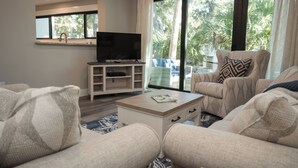 Een moderne woonkamer met een flatscreen televisie op een houten standaard, beige banken, een salontafel en een grote schuifdeur die leidt naar een balkon met uitzicht op palmbomen.