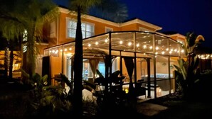Un espace extérieur bien éclairé, doté d’une structure en verre, de palmiers et d’une piscine.