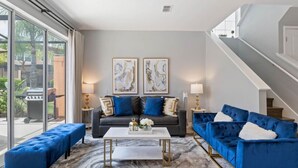 Un salon moderne avec un canapé gris, des fauteuils bleus et une table basse blanche. Deux tableaux abstraits ornent le mur, et un escalier se trouve sur la droite.