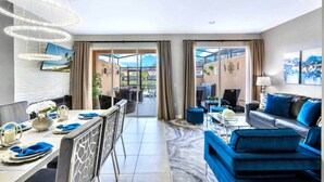 A modern hotel room with a dining area, a living room with blue sofas, and large windows offering a view of the outdoors. - #2609 Resort Home near Disney Themed Jacuzzi Pool (Kissimmee)