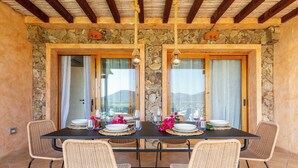 Uma área de jantar com parede de pedra, vigas de madeira e uma mesa posta para dois, com cadeiras de vime.
