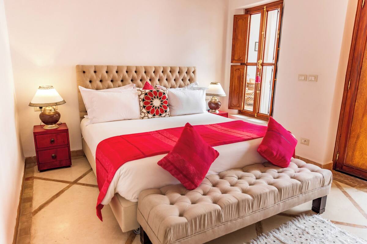 A bedroom with a tufted headboard, a large bed with white bedding and red accents, two bedside tables with lamps, and a window with wooden shutters.