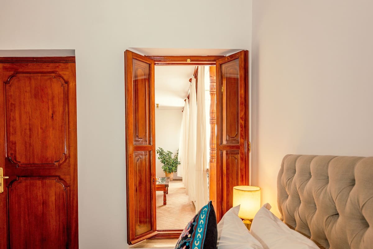 A bedroom with a bed, a bedside lamp, and a mirror reflecting a hallway.