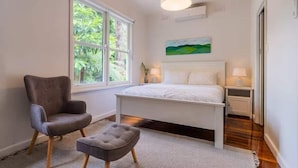 A bedroom with a bed, a chair, a nightstand, a lamp, a painting, and a window with a view of greenery. - Warburton Hideaway | MTB and Family Friendly (Warburton)