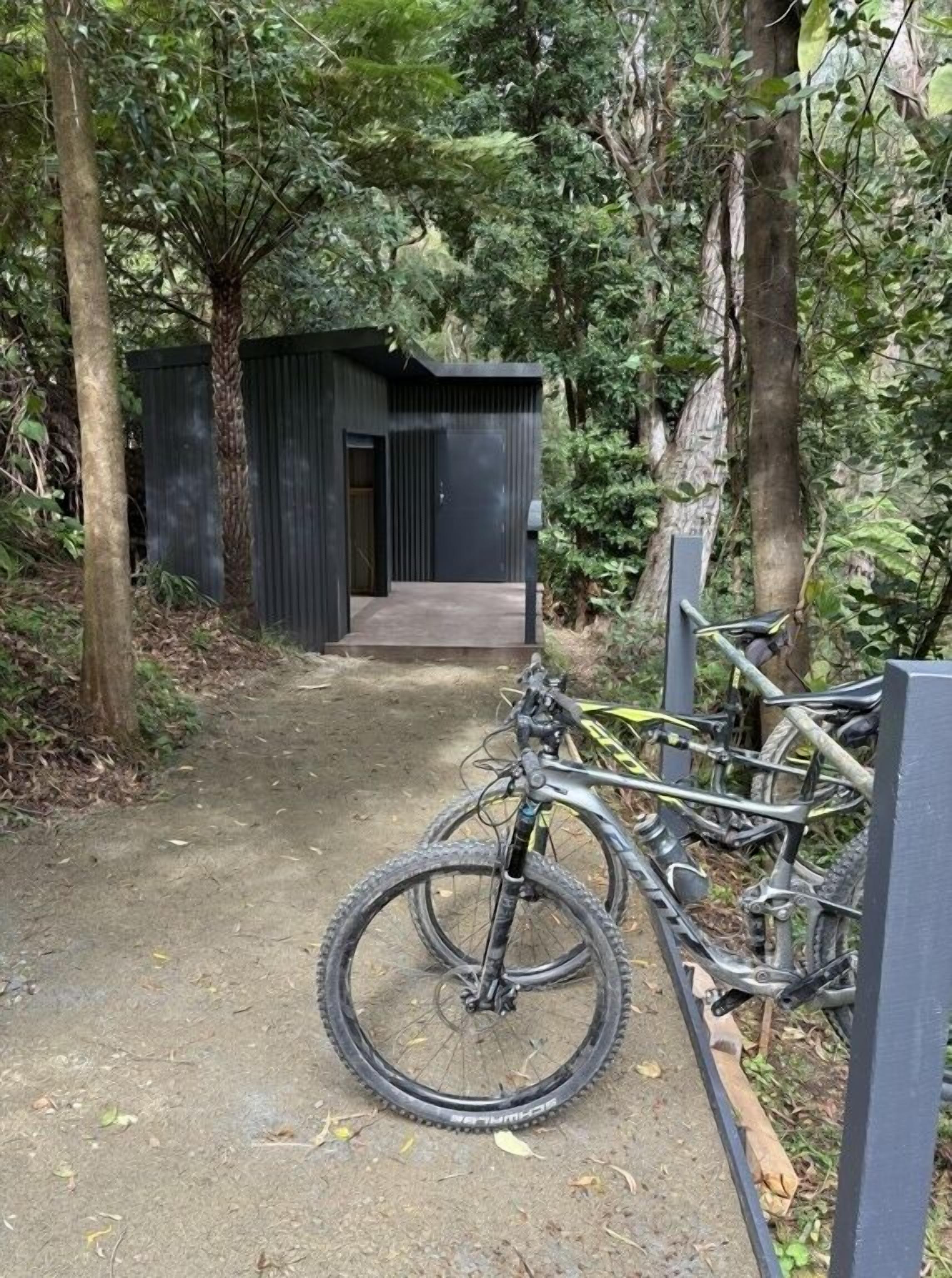 A dirt path leading to a small, dark-colored building with a door and a window. There are bicycles parked against a metal fence.