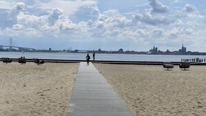 Een zandstrand met een houten looppad naar het water, een brug op de achtergrond en een stadshemelbeeld met opvallende gebouwen.