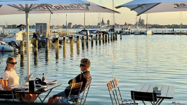 Een eetgedeelte aan het water met tafels en stoelen onder parasols, boten afgemeerd op de achtergrond en een stadszicht in de verte.