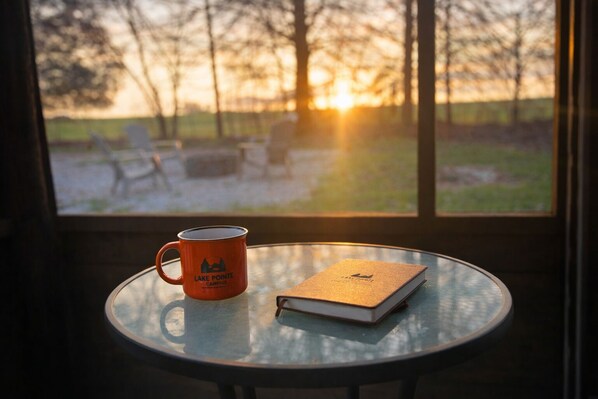 Une table sur laquelle se trouvent un livre et une tasse, le tout posé sur une table en verre, avec vue sur des arbres et un coucher de soleil à travers une fenêtre.