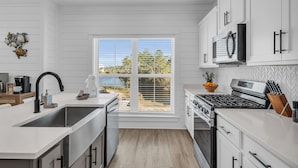 A modern kitchen with white cabinetry, a stainless steel sink, and a large window overlooking a scenic view. - Coastal Bummz | Pets | Walk to Beach & Pool (Orange Beach)