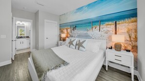 A bedroom with a beach-themed mural, a bed with starfish pillows, a nightstand with a lamp, and a view of the ocean through a window. - Coastal Bummz | Pets | Walk to Beach & Pool (Orange Beach)