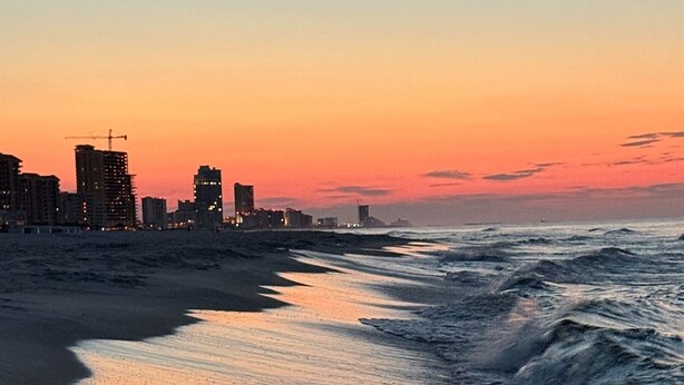 A beach with waves, a shadow of a person, and buildings in the background. - Coastal Bummz | Pets | Walk to Beach & Pool (Orange Beach)