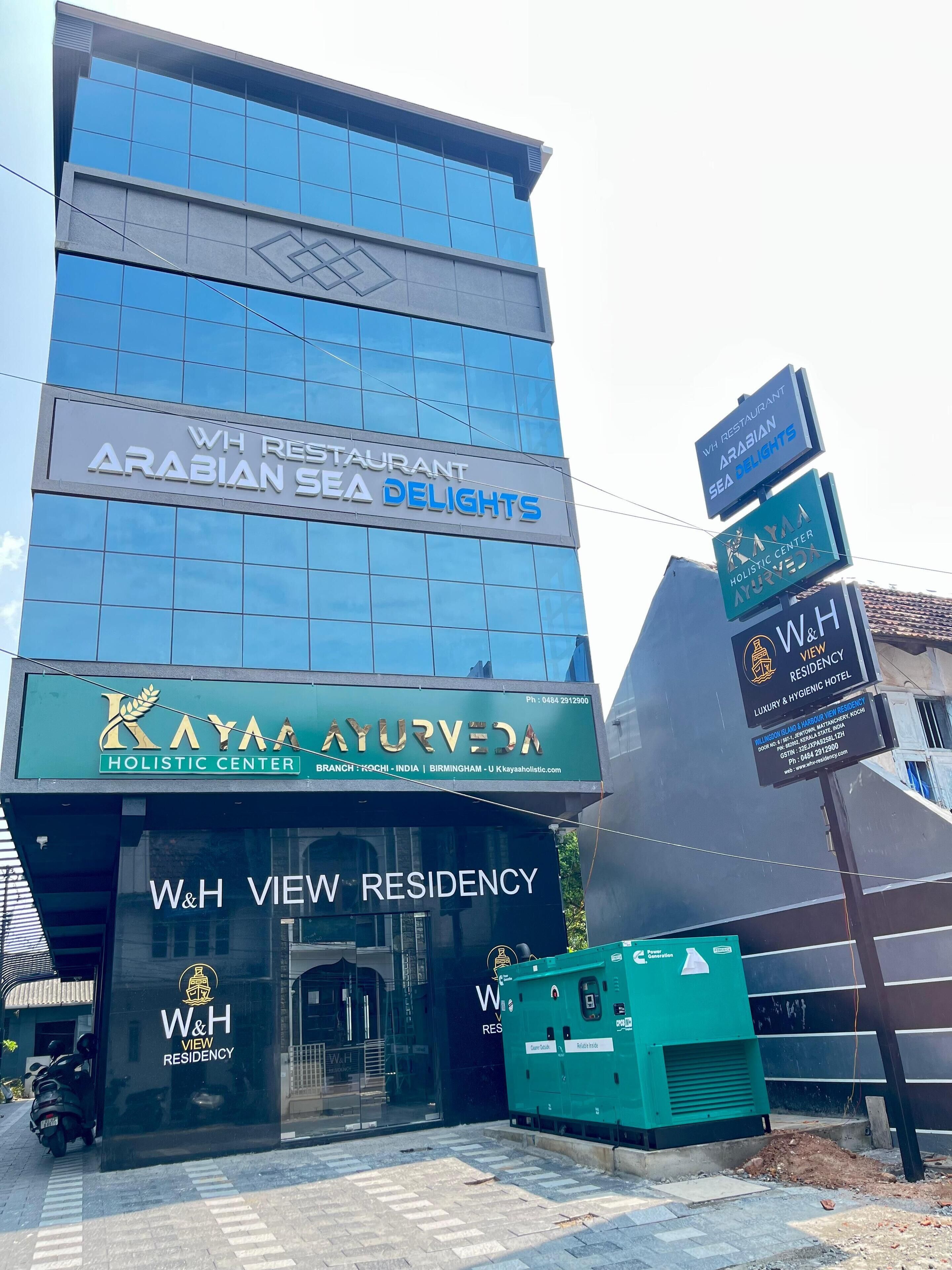 Un edificio de varios pisos con carteles para el restaurante WH, Delicias del Mar Arábigo, el centro holístico Kayaa Ayurveda y la Residencia W&H View.