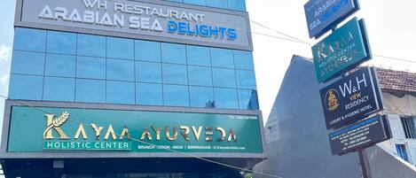 Un edificio de varios pisos con carteles para el restaurante WH, Delicias del Mar Arábigo, el centro holístico Kayaa Ayurveda y la Residencia W&H View.