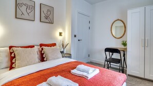 A hotel room with a bed, two pillows, a desk, a chair, a mirror, and two framed artworks on the wall. - InspoHome Bayview 1-Bed Cardiff Bay  (Cardiff)