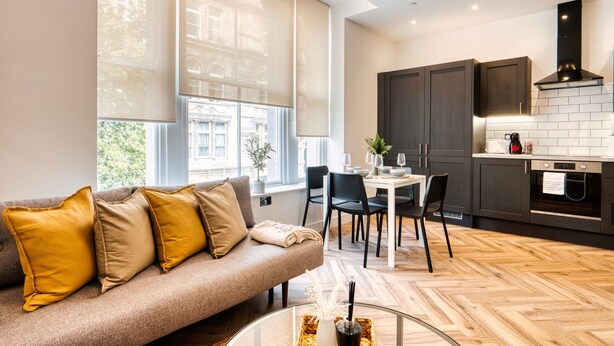 A modern living room with a sofa, dining table, and chairs. There is a kitchen area with dark cabinets and a stove. - InspoHome Bayview 1-Bed Cardiff Bay  (Cardiff)