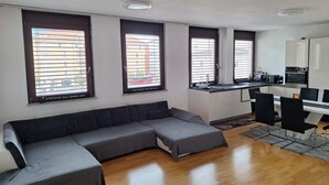 Un salon moderne avec un canapé d’angle, un coin cuisine comprenant une table et des chaises, et de grandes fenêtres équipées de stores.